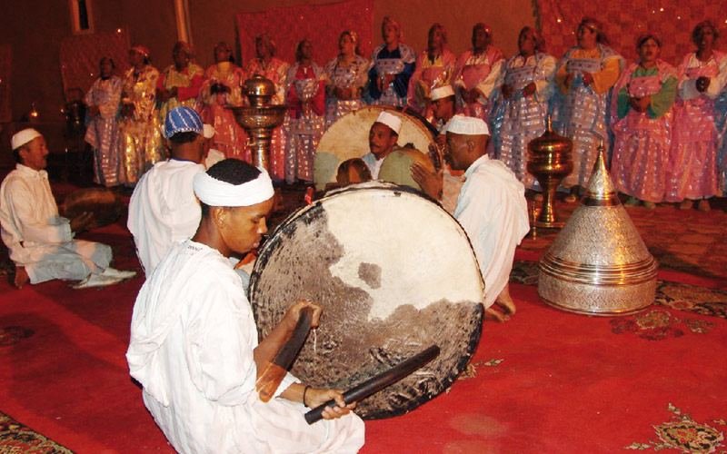 Musiciens d'Ahwach en blanc avec bendirs, femmes en robes brodées en arrière-plan, sur tapis rouges; illustration de l'article d'annonce de la 3e édition (Aujourd'hui le Maroc, 25 juillet 2014).