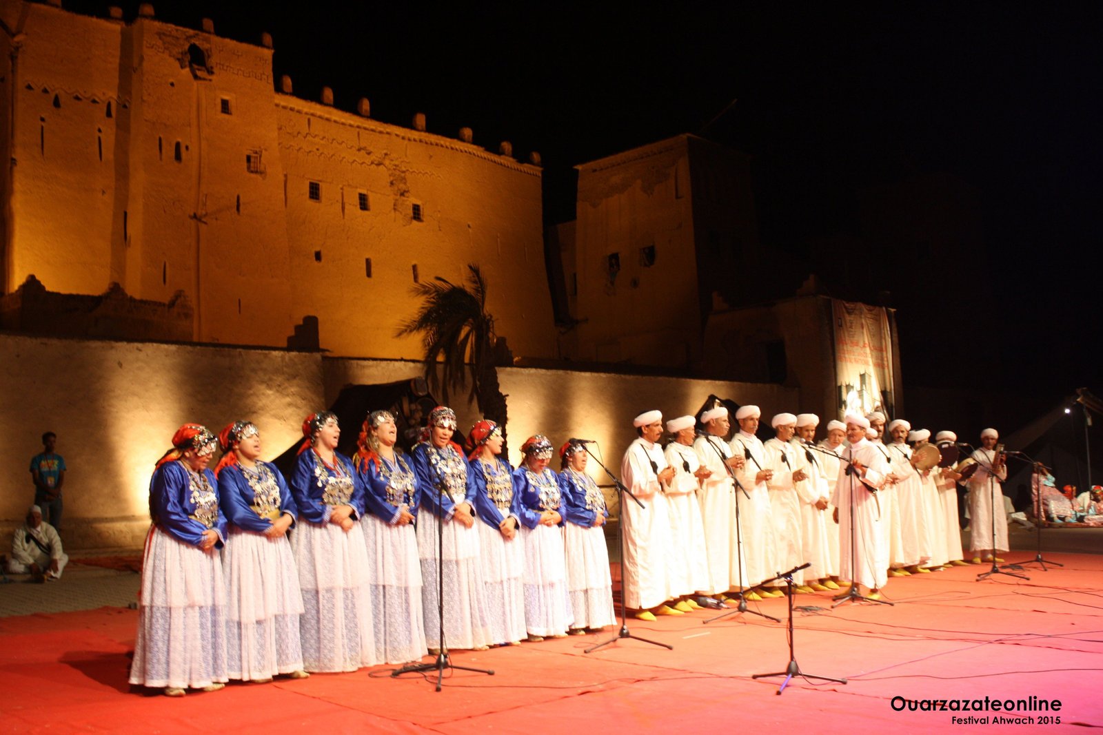 Troupe d'Ahwach sur le tapis rouge de la scène dressée au pied de la Kasbah Taourirt éclairée : rangée de femmes en caftans bleus, jupes blanches et parures d'argent, rangée d'hommes en djellabas blanches et turbans devant les micros, bannière du festival à l'arrière-plan. 4e édition, 10–12 août 2015. Filigrane « Ouarzazateonline, Festival Ahwach 2015 ».