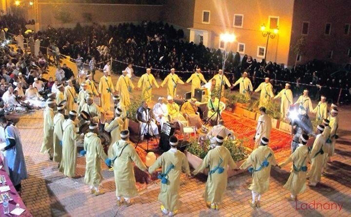 Vue plongeante d'une troupe d'Ahwach en cercle (hommes en blanc, femmes en robes vert pâle) sur tapis rouges devant un public nombreux, Ouarzazate.