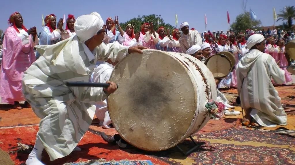 Joueur de grand tambour agenouillé en djellaba blanche, danseuses en robes roses en arrière-plan, place de la Kasbah Taourirt, ouverture de la 6e édition.