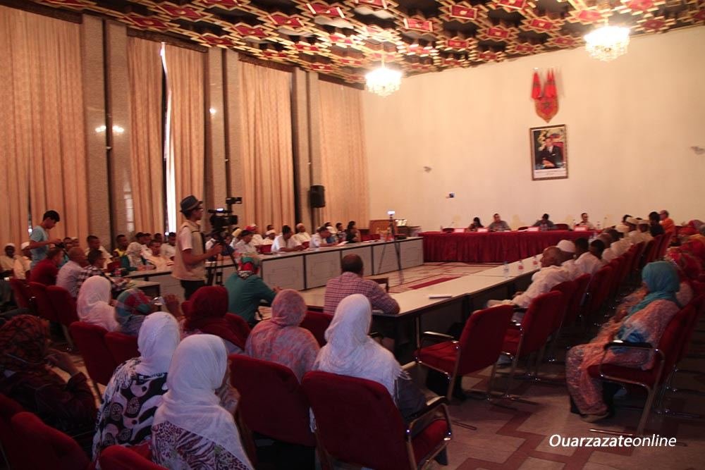 Salle du Palais des congrès de Ouarzazate pendant le colloque de la 6e édition (vendredi 11 août 2017) : public assis en fauteuils rouges, table des intervenants en U sous le portrait royal et les drapeaux, cadreur au centre. Filigrane « Ouarzazateonline ».