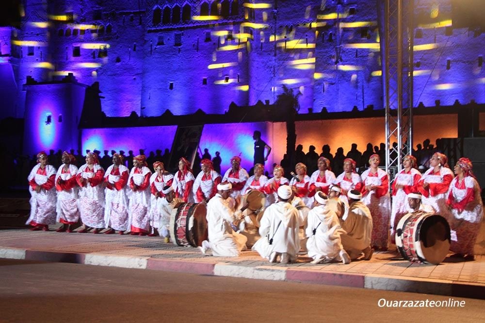 Rangée de femmes en robes blanches et rouges et foulards rouges, percussionnistes en djellabas blanches accroupis au premier plan avec tambours sur cadre et grand tambour, devant la Kasbah Taourirt éclairée en bleu et jaune, soirée du 6e Festival national des arts d'Ahwach, Ouarzazate, 10–12 août 2017. Filigrane « Ouarzazateonline ».