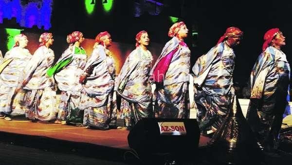 Rangée de danseuses d'Ahwach en robes argentées et foulards roses sur une scène éclairée; illustration de l'annonce de la 7e édition par Assabah.