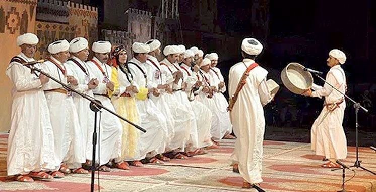 Troupe d'Ahwach en blanc, raïs au centre avec bendir, danseuses en arrière-plan, devant la façade ocre d'une kasbah; illustration de l'annonce de la 7e édition (10–12 août 2018).