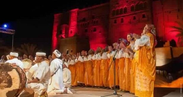Troupes d'Ahwach (femmes en robes jaunes, hommes en blanc) sur la scène au pied de la Kasbah Taourirt illuminée en rouge; illustration de l'annonce de la 8e édition (27–29 juin 2019).