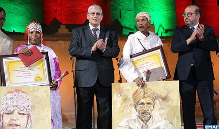 Cérémonie d'hommage sur la scène de la Kasbah Taourirt (murs éclairés en rouge et vert) : une artiste en tenue traditionnelle et un artiste en djellaba blanche et turban tiennent chacun un diplôme encadré et une rose, leurs portraits peints posés devant eux, entourés d'officiels qui applaudissent. 8e Festival national des arts d'Ahwach, Ouarzazate, 27–29 juin 2019. Filigrane « ©MAP ».