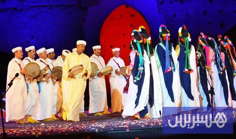 Troupe d'Ahwach (hommes en blanc avec bendirs, femmes en robes jaunes) sur une scène devant une tenture rouge; illustration de l'article du 11 juillet 2021 sur l'édition numérique.