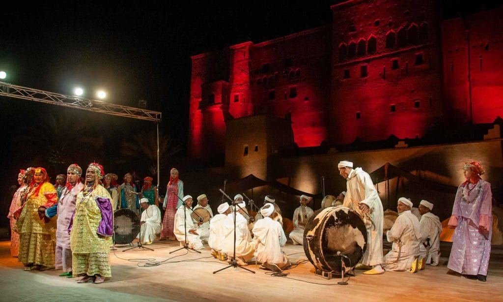Troupes d'Ahwach en robes jaunes et blanches avec bendirs sur la scène de la Kasbah Taourirt illuminée en rouge; illustration de l'annonce de la 10e édition.