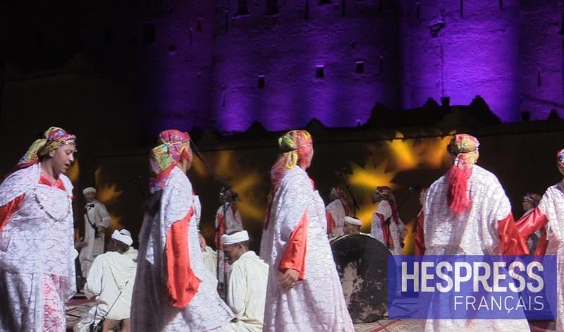 Troupe d'Ahwach de nuit : femmes en robes blanches brodées et foulards multicolores, hommes en blanc, Kasbah Taourirt éclairée en violet en arrière-plan. Photo de la MAP datée du 13 juin 2022 (identifiant MAPPH_20220613_0004), réutilisée par Hespress FR le 27 mai 2024.