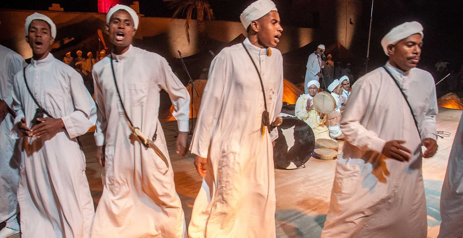 Quatre danseurs en djellabas blanches, turbans blancs et poignards en bandoulière chantent en avançant sur la scène ; derrière eux, percussionnistes assis avec tambours sur cadre et grand tambour à peau tachetée, murs de la Kasbah Taourirt éclairés. Photographie de la médiathèque du ministère de la Jeunesse, de la Culture et de la Communication (fichier « Ahwach », téléversé le 10 mars 2022), édition photographiée non précisée.