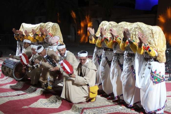 Soirée d'ouverture : troupe féminine en robes blanches et bijoux d'argent, musiciens assis au premier plan, scène nocturne, 12 mai 2023.