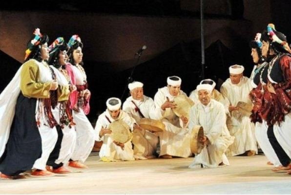 Troupe d'Ahwach sur scène : femmes en robes blanches brodées et bijoux d'argent, hommes en blanc accroupis avec bendirs; article sur l'ouverture de la 12e édition.