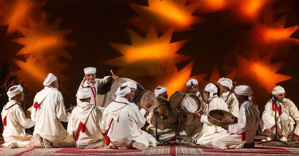 Troupe d'Ahwach assise et debout sur une scène avec projection d'étoiles orange en fond; illustration de l'annonce de la 12e édition (14 mai 2024).