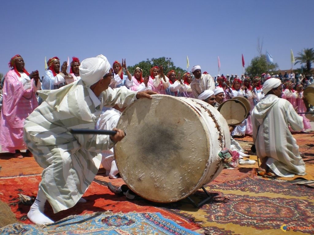 Joueur de grand tambour (ganga) en djellaba blanche au premier plan, troupe en rose et blanc avec drapeaux en arrière-plan, en plein jour sur tapis; illustration de la dépêche MAP sur la deuxième journée.