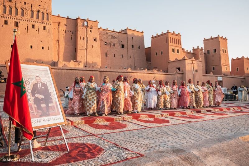 Troupe d'Ahwach en robes colorées alignée sur tapis rouges devant la Kasbah Taourirt, drapeau marocain et portrait royal sur chevalet; photo d'illustration de l'annonce de la 14e édition (6 octobre 2026).